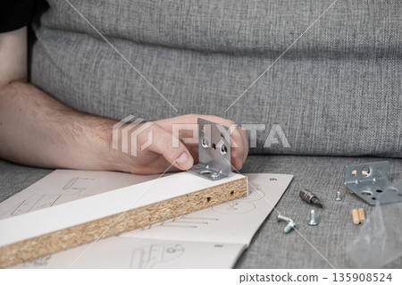 Man's hands assembling flat-pack furniture, attaching metal bracket to particle board piece, carefully following instruction manual for home DIY project Man's hands assembling flat-pack furniture, attaching metal bracket to particle board piece, carefully following instruction manual for home DIY project 135908524