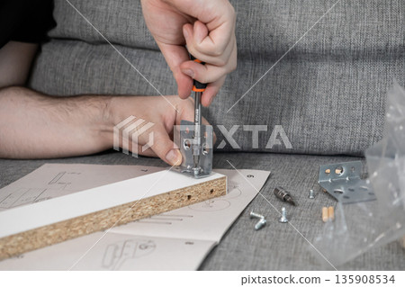 Adult hands meticulously using a screwdriver to fasten a metal bracket onto a particle board, illustrating home furniture assembly and practical do-it-yourself projects 135908534