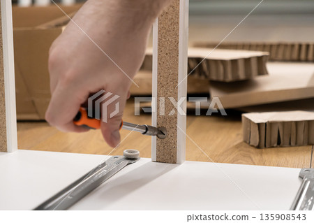 Close-up of a hand using a screwdriver to assemble white flat-pack furniture, representing home improvement, practical skills, and DIY project setup 135908543