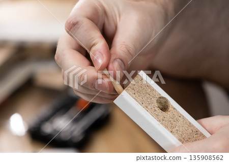 Person's hand carefully inserting a wooden dowel into a pre-drilled hole of a particleboard panel, showcasing home furniture assembly and practical DIY 135908562