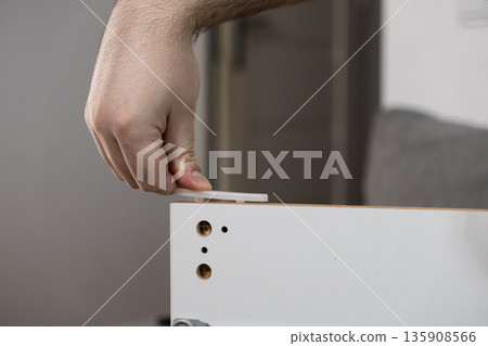 Adult hand meticulously inserting a white plastic dowel pin into a wooden furniture panel, symbolizing DIY assembly and home improvement work Adult hand meticulously inserting a white plastic dowel pin into a wooden furniture panel, symbolizing DIY assembly and home improvement work 135908566