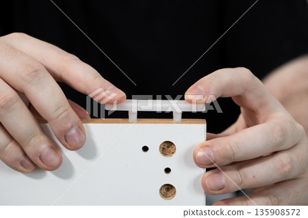 Hands carefully assembling new white flat-pack furniture, positioning a plastic connector onto wooden dowels, symbolizing modern home improvement and DIY assembly effort 135908572