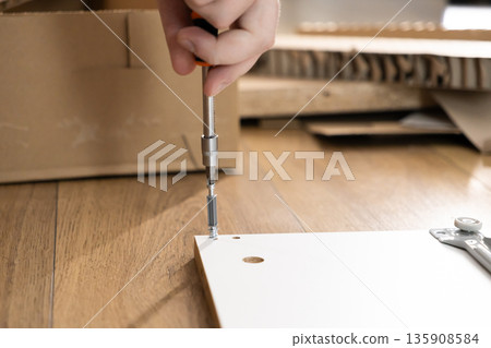Close-up of a person's hand meticulously using a screwdriver to assemble a white furniture board, symbolizing hands-on home improvement and DIY project completion Close-up of a person's hand meticulously using a screwdriver to assemble a white furniture board, symbolizing hands-on home improvement and DIY project completion 135908584