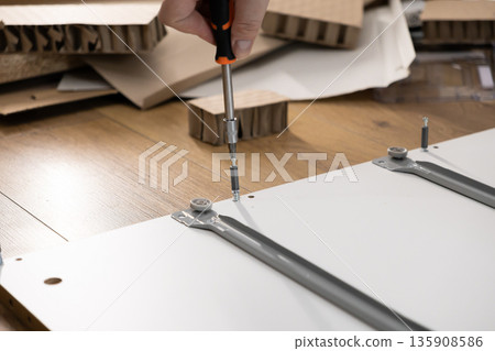 Persons hand using a screwdriver to precisely fasten components of modern flat-pack furniture, representing do-it-yourself home assembly and renovation projects 135908586