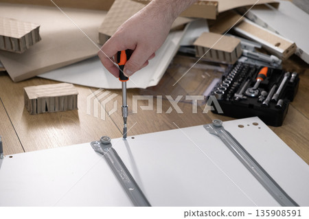Close-up of a person's hand using a screwdriver to assemble new white flat-pack furniture, illustrating a practical DIY home improvement project and modern craftsmanship skill Close-up of a person's hand using a screwdriver to assemble new white flat-pack furniture, illustrating a practical DIY home improvement project and modern craftsmanship skill 135908591