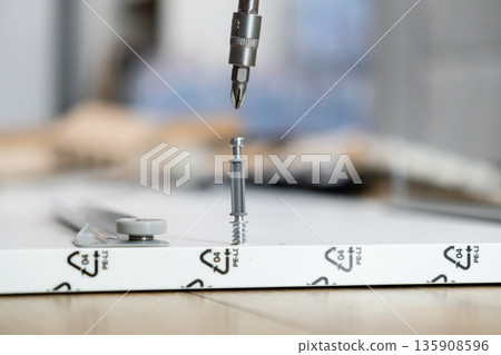 Screwdriver bit aligning to fasten a furniture connector screw into a white flat-pack board, symbolizing DIY assembly and home improvement project 135908596