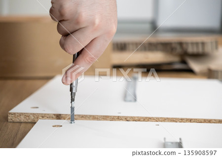 Person's hand uses a screwdriver to assemble new flat-pack furniture, illustrating DIY home improvement, renovation, and practical skill development 135908597