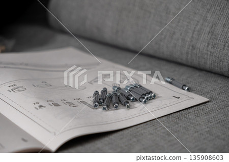 Detailed furniture assembly instruction manual with various screws and fasteners placed on a grey sofa, illustrating a practical home improvement project Detailed furniture assembly instruction manual with various screws and fasteners placed on a grey sofa, illustrating a practical home improvement project 135908603