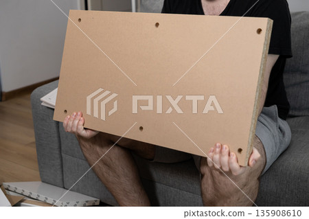 Man holding a brown wood panel with pre-drilled holes, preparing for home furniture assembly, symbolizing a personal do-it-yourself project and practical home improvement 135908610