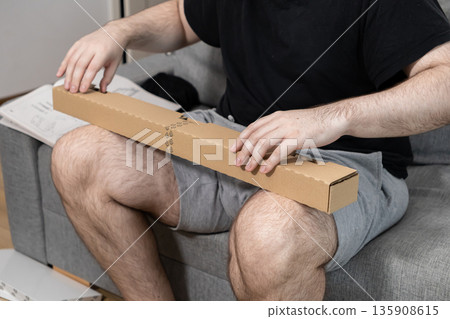 Man in comfortable clothing carefully unboxing a long brown cardboard package, symbolizing modern online shopping, home delivery, and furniture self-assembly 135908615