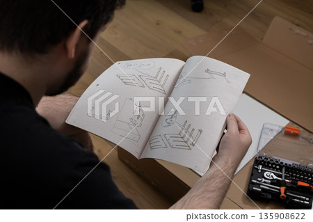Young adult carefully reading detailed furniture assembly guide for a new purchase, focused on successful DIY home improvement project setup Young adult carefully reading detailed furniture assembly guide for a new purchase, focused on successful DIY home improvement project setup 135908622