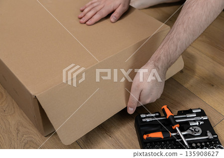Man's hands carefully opening a large brown cardboard box on a wooden floor, with a toolkit ready for home assembly, symbolizing a new DIY project 135908627