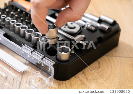 Hand selecting a chrome metal socket from a well-organized professional tool kit, highlighting precision, readiness, and practical home repair or industrial maintenance Hand selecting a chrome metal socket from a well-organized professional tool kit, highlighting precision, readiness, and practical home repair or industrial maintenance 135908630