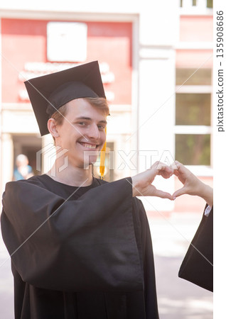 Young Caucasian man in graduation gown showing heart sign.  135908686