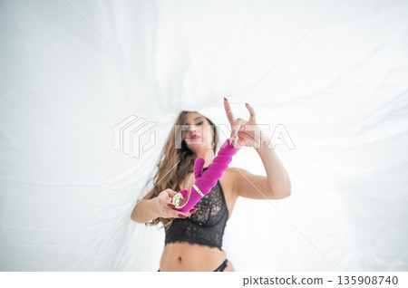 A woman in lingerie sits under a sheet and holds a vibrator. A woman in lingerie sits under a sheet and holds a vibrator. 135908740