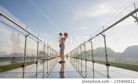 Caucasian woman holds her little son in her arms while standing on a glass bridge in the mountains. Caucasian woman holds her little son in her arms while standing on a glass bridge in the mountains. 135908811