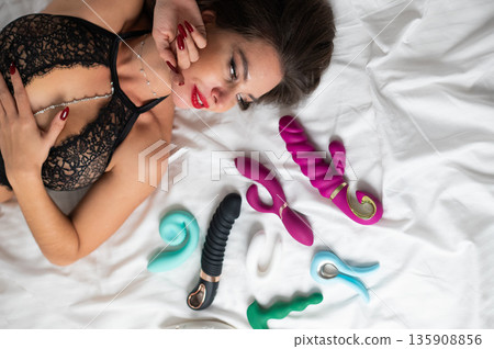 A beautiful Caucasian woman in black lingerie lies among sex toys.  135908856