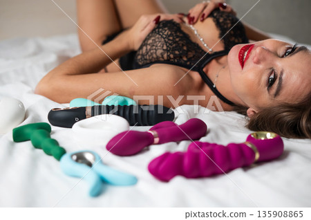 A beautiful Caucasian woman in black lingerie lies among sex toys.  135908865