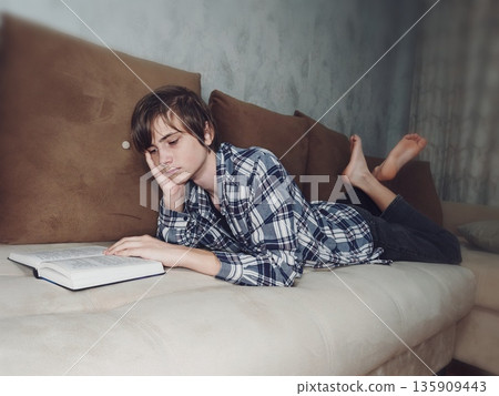 Teenage Boy Reading Book on Couch Indoors. Young boy lying on brown couch, focused reading open book in cozy home interior. Teenage Boy Reading Book on Couch Indoors. Young boy lying on brown couch, focused reading open book in cozy home interior. 135909443