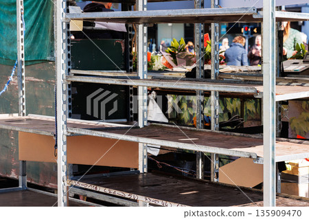 Empty shelves of metal rack with at an outdoor market stall, with flower crates and people blurred in the background. 135909470
