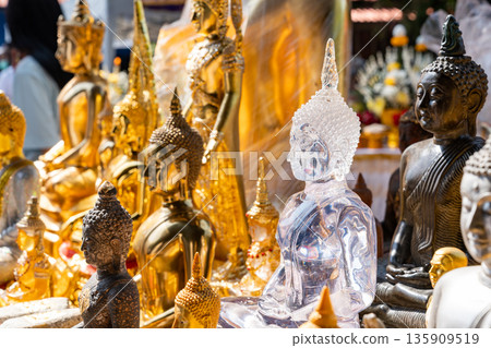 Transparent glass buddha statue in meditation posture among various buddha statues on the blurry background of people and buildings in Thailand temple. Transparent glass buddha statue in meditation posture among various buddha statues on the blurry background of people and buildings in Thailand temple. 135909519