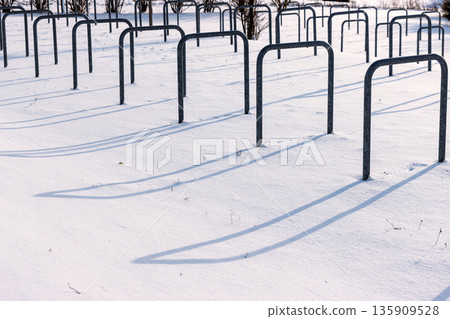 Snow covered bicycle parking area with empty metal bike rack casting long shadows in bright winter sunlight. Snow covered bicycle parking area with empty metal bike rack casting long shadows in bright winter sunlight. 135909528