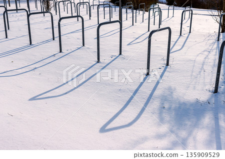 Snow covered bicycle parking area with empty metal bike rack casting long shadows in bright winter sunlight. Snow covered bicycle parking area with empty metal bike rack casting long shadows in bright winter sunlight. 135909529