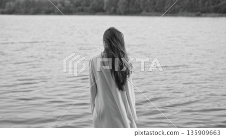 Lonely woman with long hair standing facing water, symbolizing burnout, depression, midlife crisis, emotional fatigue, and introspective solitude in monochrome. 135909663
