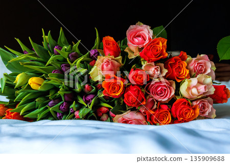 Romantic mixed bouquet of roses and tulips lying on a bed blanket in a bedroom, with a dark black background. Romantic mixed bouquet of roses and tulips lying on a bed blanket in a bedroom, with a dark black background. 135909688