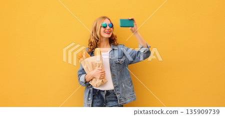 Happy smiling young woman taking selfie with smartphone holds grocery shopping paper bag with bread 135909739