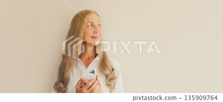 Happy smiling woman with phone in white shirt, girl holding smartphone looking away on background Happy smiling woman with phone in white shirt, girl holding smartphone looking away on background 135909764
