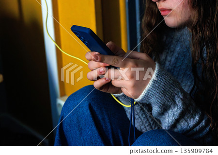 Close up of young woman hands holding and using smartphone with charging cable in the hotel room Close up of young woman hands holding and using smartphone with charging cable in the hotel room 135909784