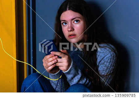 Thoughtful young woman looking at camera while holding smartphone with charging cable indoors 135909785