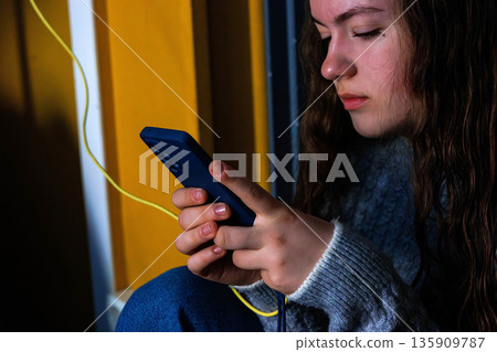 Young woman holding and using smartphone with charging cable indoors 135909787