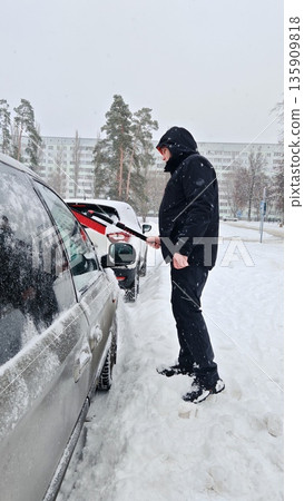 Man Cleaning Snow from Car in Winter. Person using snow brush to clean snow off beige minivan during heavy snowfall in city yard. Man Cleaning Snow from Car in Winter. Person using snow brush to clean snow off beige minivan during heavy snowfall in city yard. 135909818