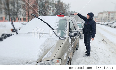 Man Cleaning Snow from Car in Winter. Person using snow brush to clean snow off beige minivan during heavy snowfall in city yard. Man Cleaning Snow from Car in Winter. Person using snow brush to clean snow off beige minivan during heavy snowfall in city yard. 135910043