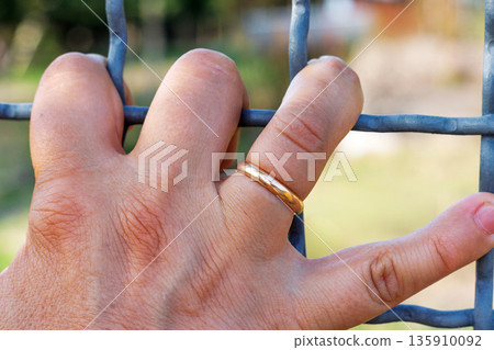 Close up of a human hand gripping a metal wire fence, wearing a gold wedding ring Close up of a human hand gripping a metal wire fence, wearing a gold wedding ring 135910092