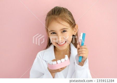 Smiling child dressed as dentist holding toothbrush and teeth model. Concept of dental education, oral hygiene awareness, prevention and International Dentist Day. Smiling child dressed as dentist holding toothbrush and teeth model. Concept of dental education, oral hygiene awareness, prevention and International Dentist Day. 135911284