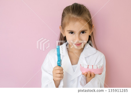 Smiling child in medical coat holding toothbrush and dental model. Concept of oral hygiene education, prevention, pediatric dentistry and healthy habits for children. Smiling child in medical coat holding toothbrush and dental model. Concept of oral hygiene education, prevention, pediatric dentistry and healthy habits for children. 135911285
