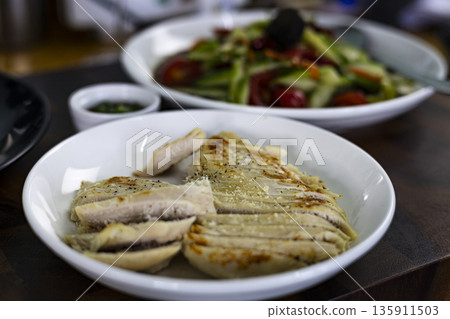 Grilled Chicken Breast On White Ceramic, A clean photo of grilled chicken breast , golden char marks and fresh herbs in bright natural light, for healthy meal or simple elegance documentary. Grilled Chicken Breast On White Ceramic, A clean photo of grilled chicken breast , golden char marks and fresh herbs in bright natural light, for healthy meal or simple elegance documentary. 135911503