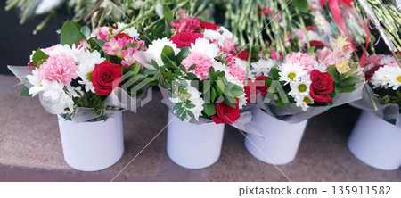 Several vibrant bouquets featuring red roses, pink carnations, white daisies, and alstroemeria are displayed in matching white cylindrical vases outdoors Several vibrant bouquets featuring red roses, pink carnations, white daisies, and alstroemeria are displayed in matching white cylindrical vases outdoors 135911582
