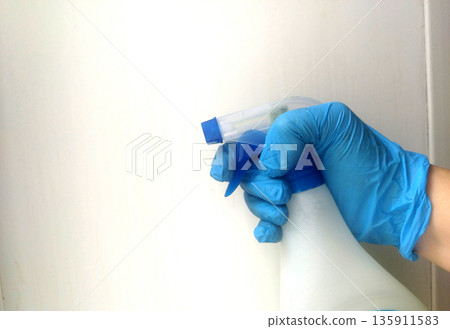 Hand wearing a bright blue disposable glove firmly grips the trigger mechanism of a white spray bottle against a simple, light background, ready for cleaning tasks Hand wearing a bright blue disposable glove firmly grips the trigger mechanism of a white spray bottle against a simple, light background, ready for cleaning tasks 135911583