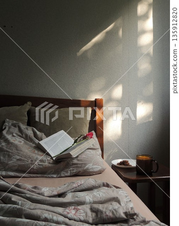 A vertical photo of a bedroom with a wooden bed and beige beautiful cozy bedding, with an open book and a flower on the pillow and a mug of tea and homemade cookies on a stand, in a room with sun 135912820