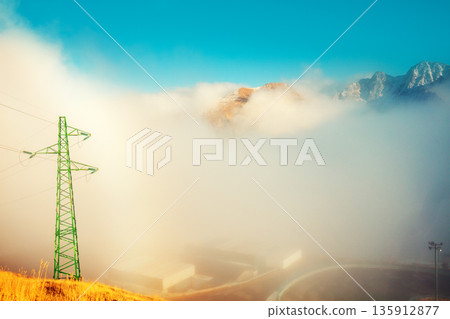 Mountains in clouds. Alpine winding highway above the clouds. Power transmission line in the mountains. Transmission facilities at sunset. Pyrenees, Andorra, Europe Mountains in clouds. Alpine winding highway above the clouds. Power transmission line in the mountains. Transmission facilities at sunset. Pyrenees, Andorra, Europe 135912877