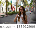 Smiling woman holding sunglasses on sunny city street 135912909