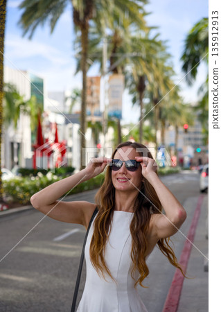 Stylish woman adjusting sunglasses on sunny city street 135912913