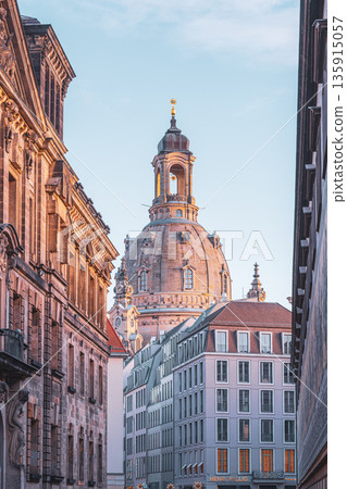 Frauenkirche stands tall in Dresden. The view captures the church's dome rising above nearby buildings. It is early morning, and the sky is clear. Visitors explore the area and take photos. 135915057