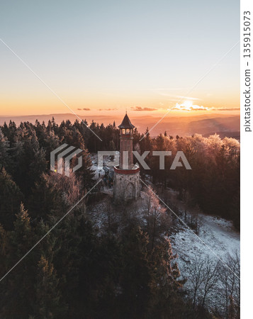 Stepanka lookout tower nestled among the trees of the Jizera Mountains during winter. The sunset casts warm colors over the snow-covered landscape. Stepanka lookout tower nestled among the trees of the Jizera Mountains during winter. The sunset casts warm colors over the snow-covered landscape. 135915073