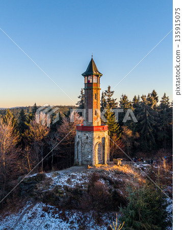 Stepanka lookout tower stands tall in the Jizera Mountains. The sunset light casts shadows on the snow-covered ground. Trees surround the tower, creating a beautiful landscape. Stepanka lookout tower stands tall in the Jizera Mountains. The sunset light casts shadows on the snow-covered ground. Trees surround the tower, creating a beautiful landscape. 135915075