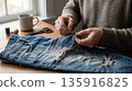 Hands mend ripped jeans with needle. Jeans repair and jeans upcycling on wooden table with thread, scissors, coffee mug, window light, for earth day, fashion revolution week, slow fashion concept 135916825